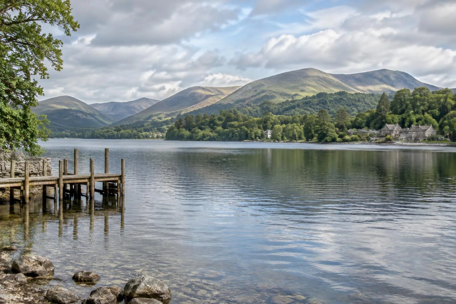 Lake Windermere with a wooden lakeside pier, stone cottages and green hills beneath a cloudy British sky